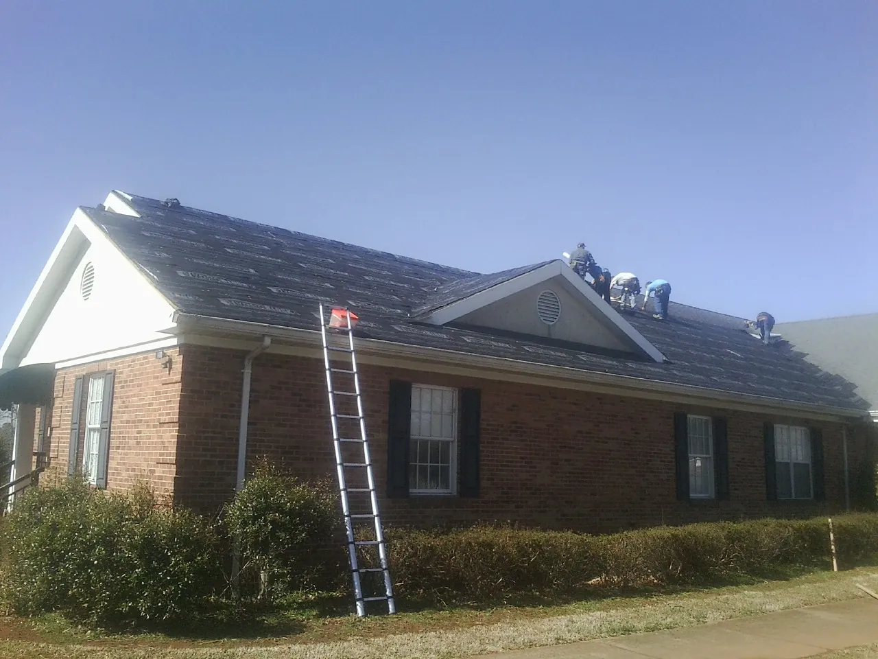 Crew tearing shingles off a brick ranch roof with a ladder set — Emergency Roof Repair work by our crew in Northville