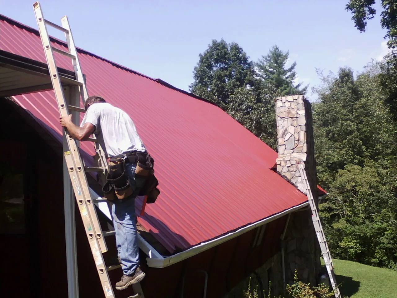 Roofer on a ladder beside a red metal roof and stone chimney — Soffit & Fascia Repair by Roofing Brothers in Northville